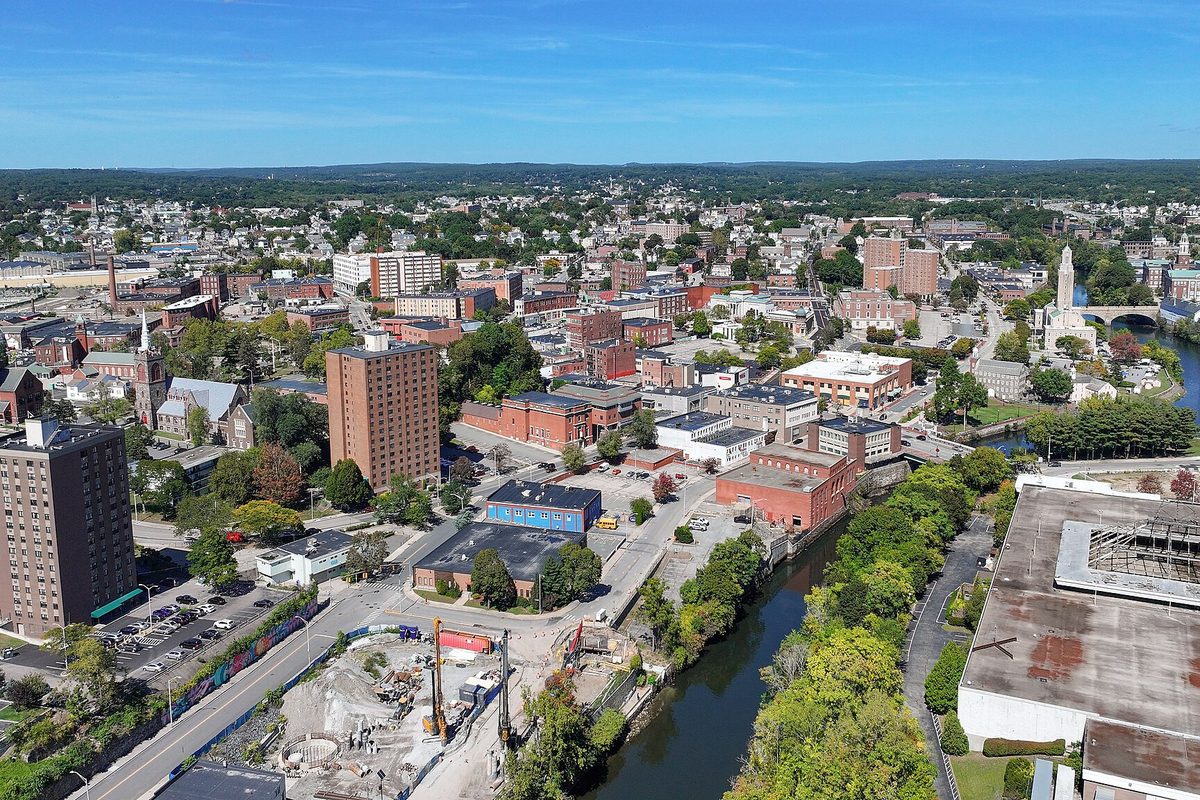 Aerial view of downtown Pawtucket with brick mill buildings, a tall brick apartment tower, and the river curving past a construction site and parking lots