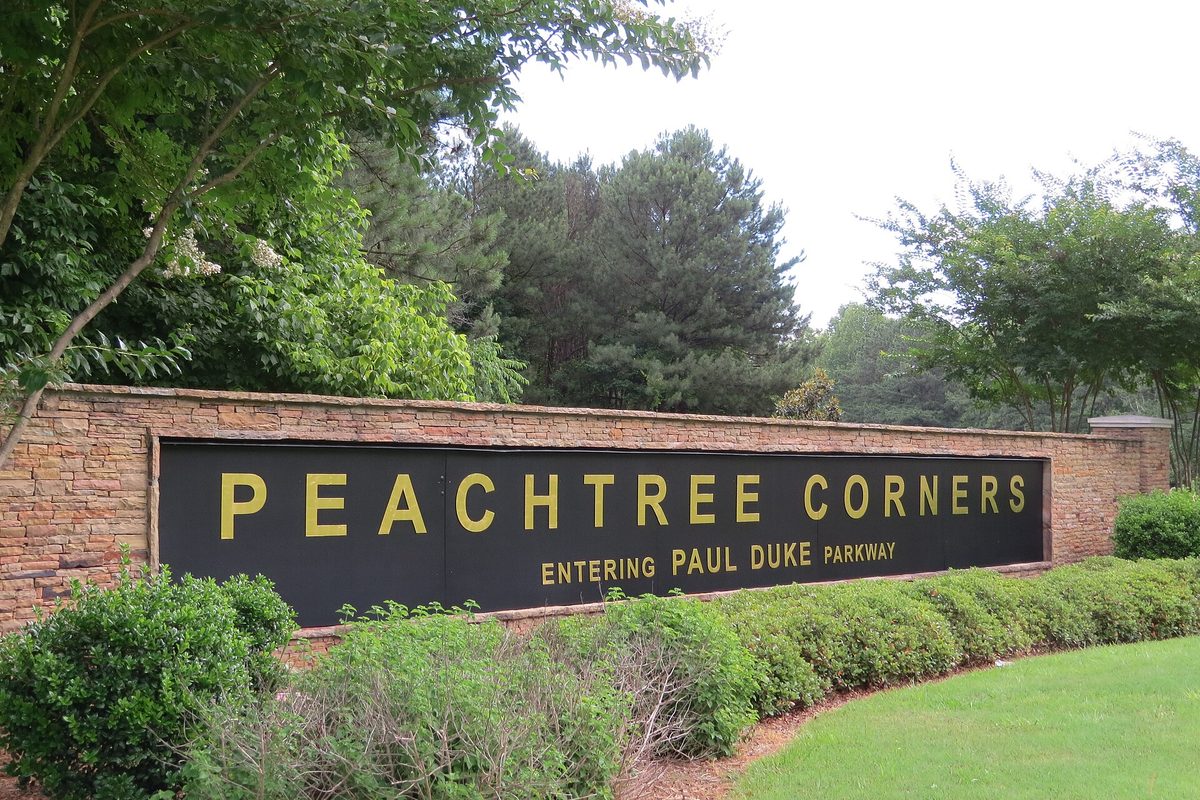 A stone wall sign with gold letters reading Peachtree Corners, Entering Paul Duke Parkway, set among shrubs and tall trees
