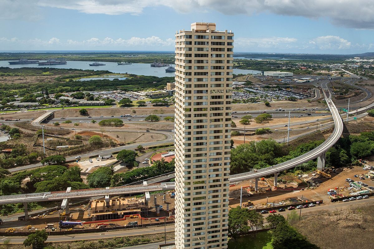 A tall condominium tower in Pearl City with the elevated Honolulu rail line curving past it and Pearl Harbor, with ships at anchor, in the distance