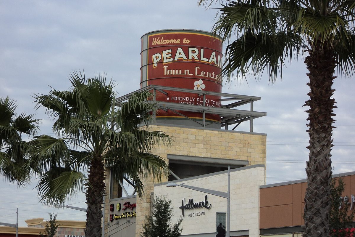 A red water-tower-style sign reading Welcome to Pearland Town Center on a stone tower, framed by palm trees above the shopping center's storefronts