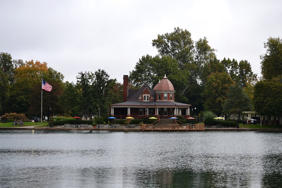 A red-roofed house with a domed corner tower and a wraparound porch on the shore of a lake in a Pekin park, with a U.S. flag and bright umbrellas