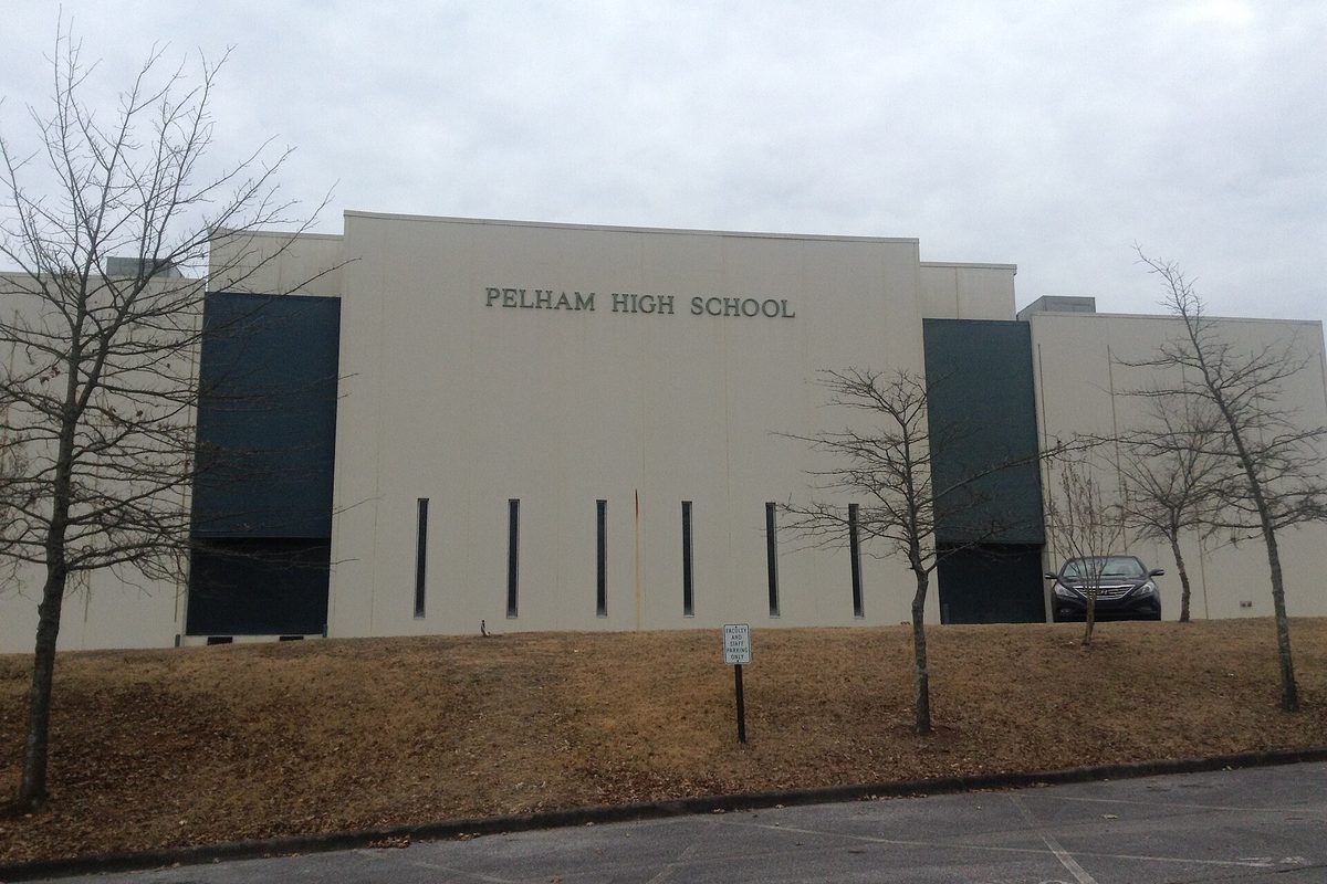 The pale front wall of Pelham High School with its name in raised letters, tall dark window panels and bare winter trees on a straw-covered slope