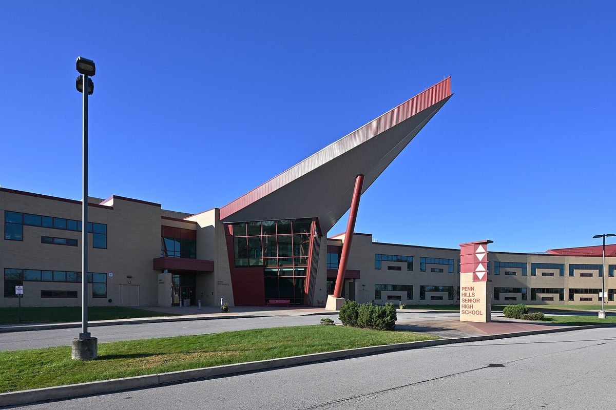 Penn Hills Senior High School, a tan building with a red-trimmed roof that angles up over a glass entrance, a school sign and lawn in front under a clear blue sky