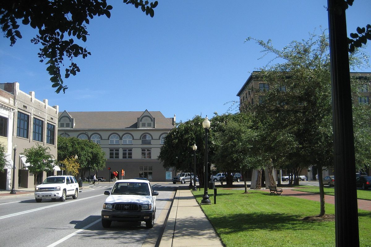 A white pickup driving down a downtown Pensacola street lined with historic brick buildings, street lamps and a grassy median under a blue sky