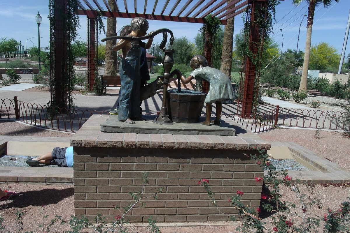 Bronze statue of two children working a hand water pump on a brick base under a red pergola in Peoria's Osuna Park, site of the 1889 town well