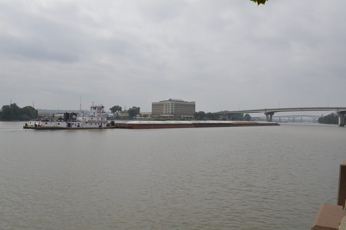 A white towboat pushing a long line of barges along the Illinois River at Peoria under a grey sky, with a highway bridge and a riverside office building behind