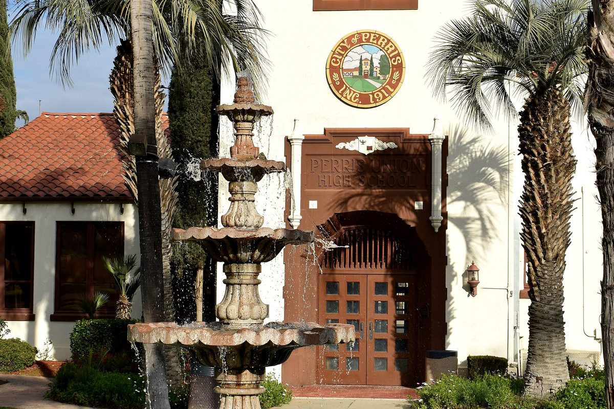 A tiered stone fountain in front of Perris City Hall, the former Perris Union High School, with arched wooden doors, palm trees and the city seal