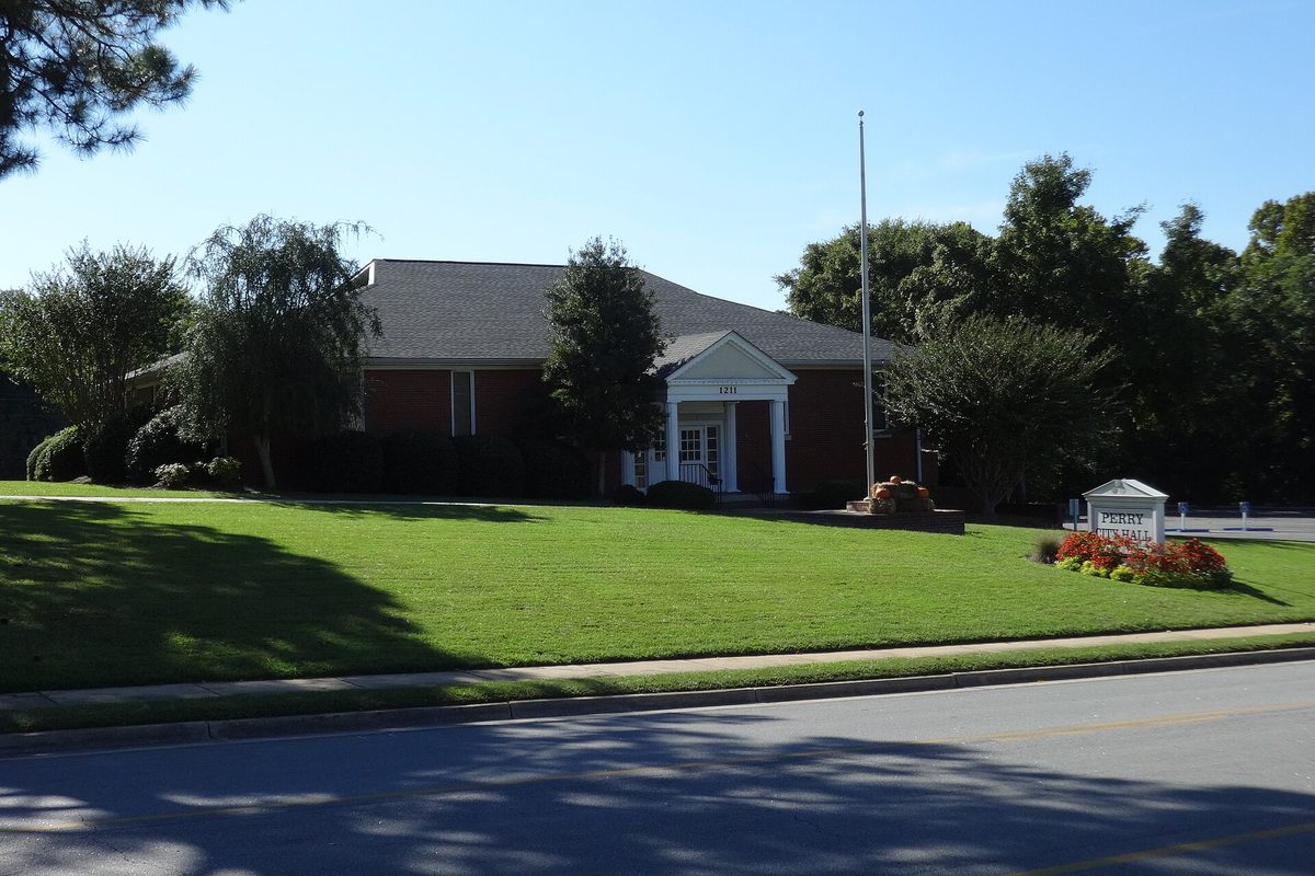 Perry City Hall, a one-story red brick building with a white columned entrance, a flagpole and a flower bed by its sign, behind a wide green lawn