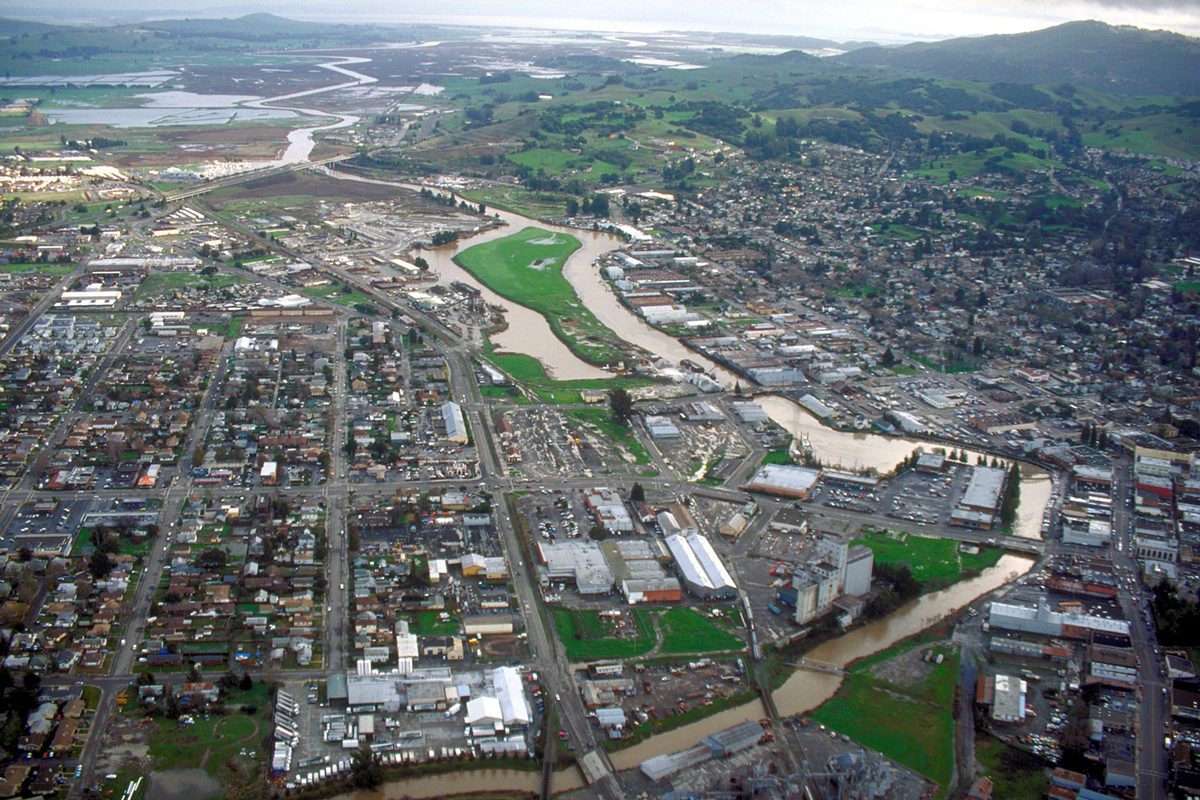 Aerial view of Petaluma with the brown Petaluma River curving through the middle of the city and green hills and the bay in the distance