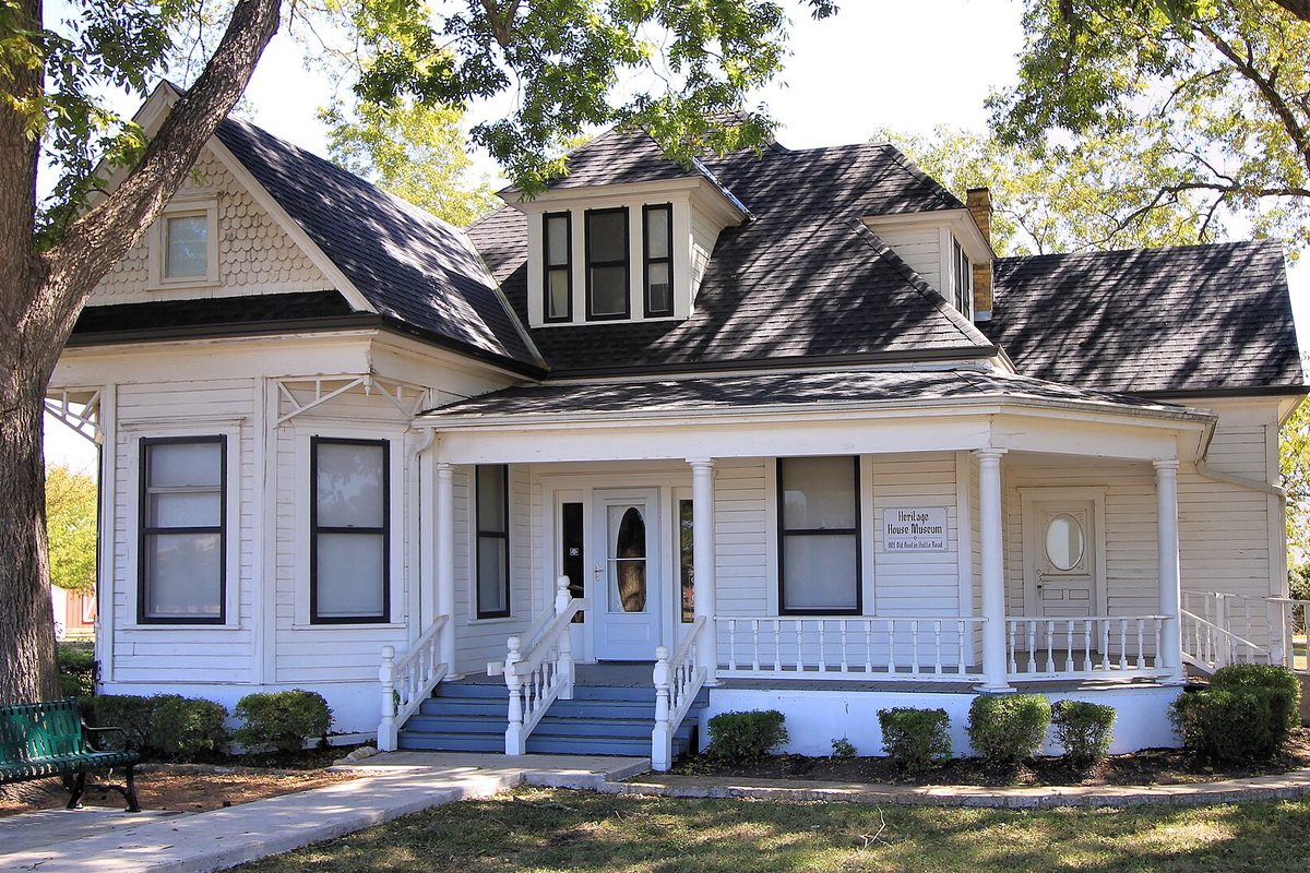 The white 1913 Bohls House, now the Heritage House Museum, with its wide front porch and dark shingle roof under shade trees in Pflugerville