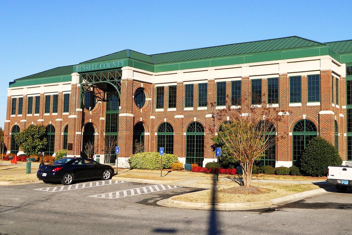 The red-brick Russell County Courthouse in Phenix City with a green metal roof and arched windows, a black car parked out front on a sunny day