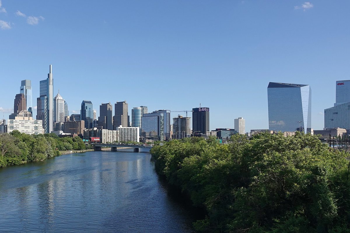 The Philadelphia skyline seen up the Schuylkill River, with towers on the left, a glass office block on the right and thick green trees along both banks