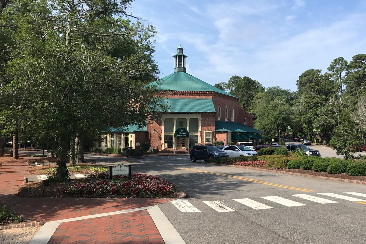 The Theatre in Pinehurst, a brick building with a green roof and white cupola, beside a crosswalk and tall trees on a sunny day