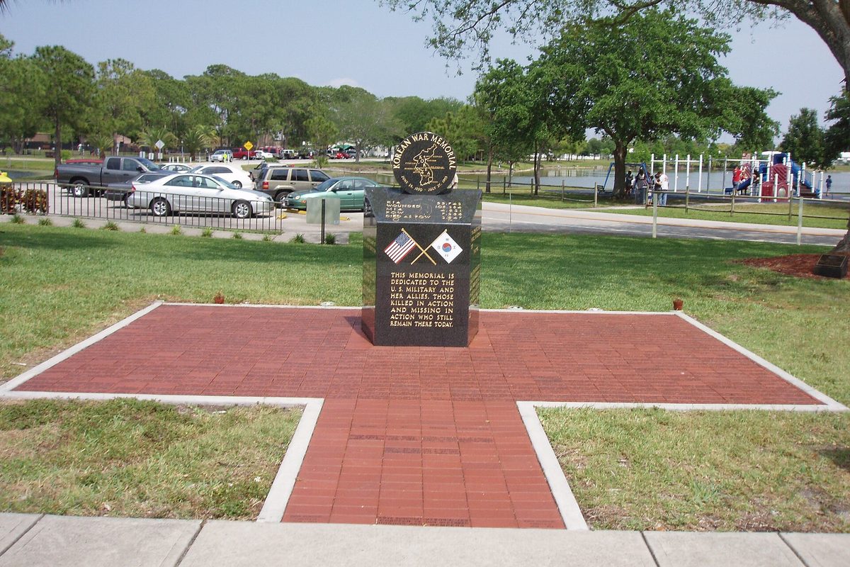 The Korean War memorial in Pinellas Park, a black stone marker on a red brick cross-shaped plaza, with a playground, a lake and parked cars behind it