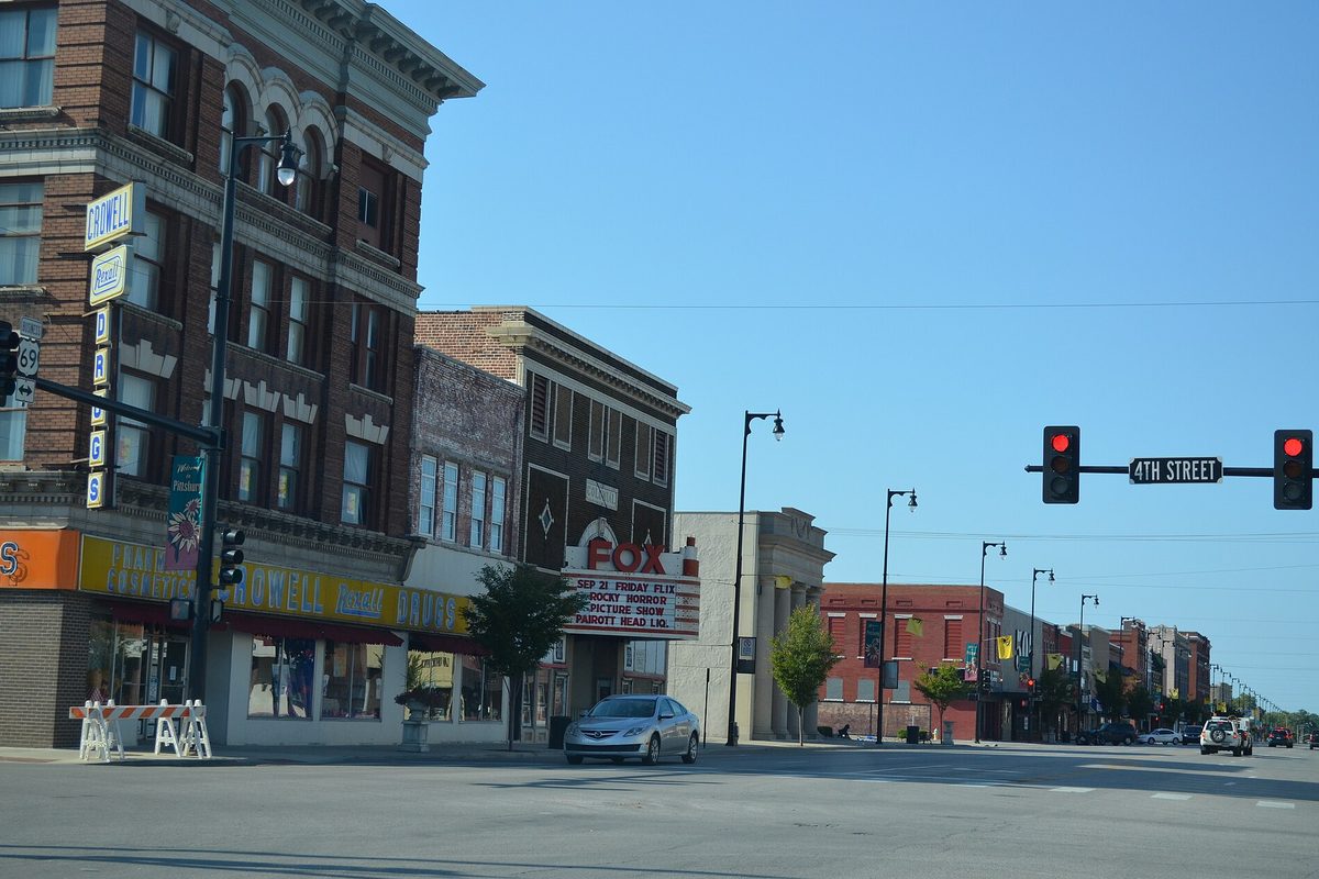 Downtown Pittsburg at the 4th Street traffic lights, with the Colonial Fox Theater marquee and old brick storefronts under a clear blue sky