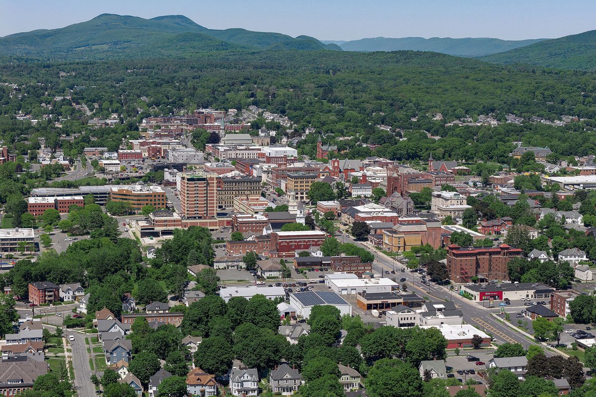 Aerial view of downtown Pittsfield, brick and stone buildings set among green trees, with wooded mountains rising behind the city