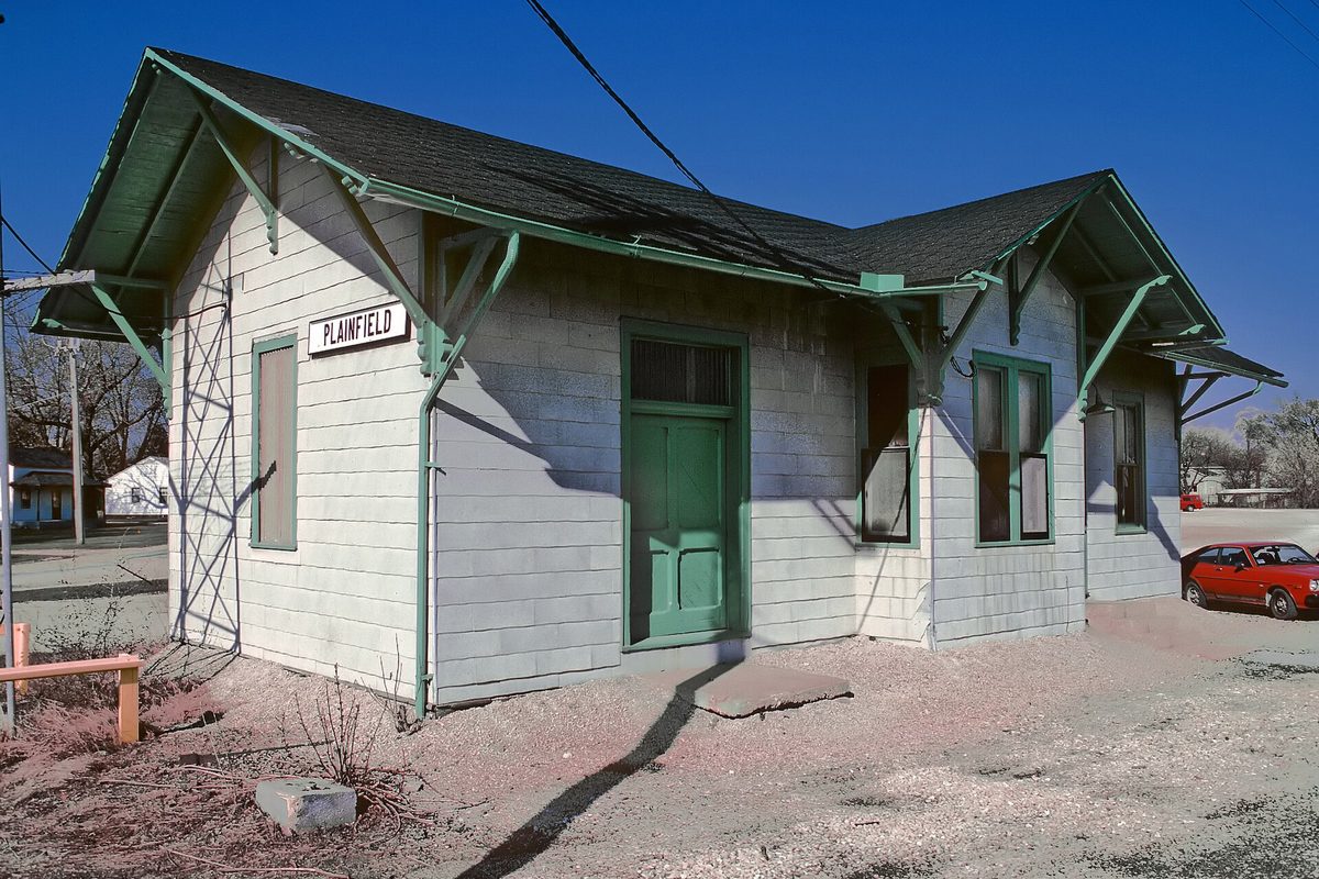 The old Plainfield railroad depot, white siding with green trim, doors and a Plainfield sign, under a deep blue sky with a red car parked beside it