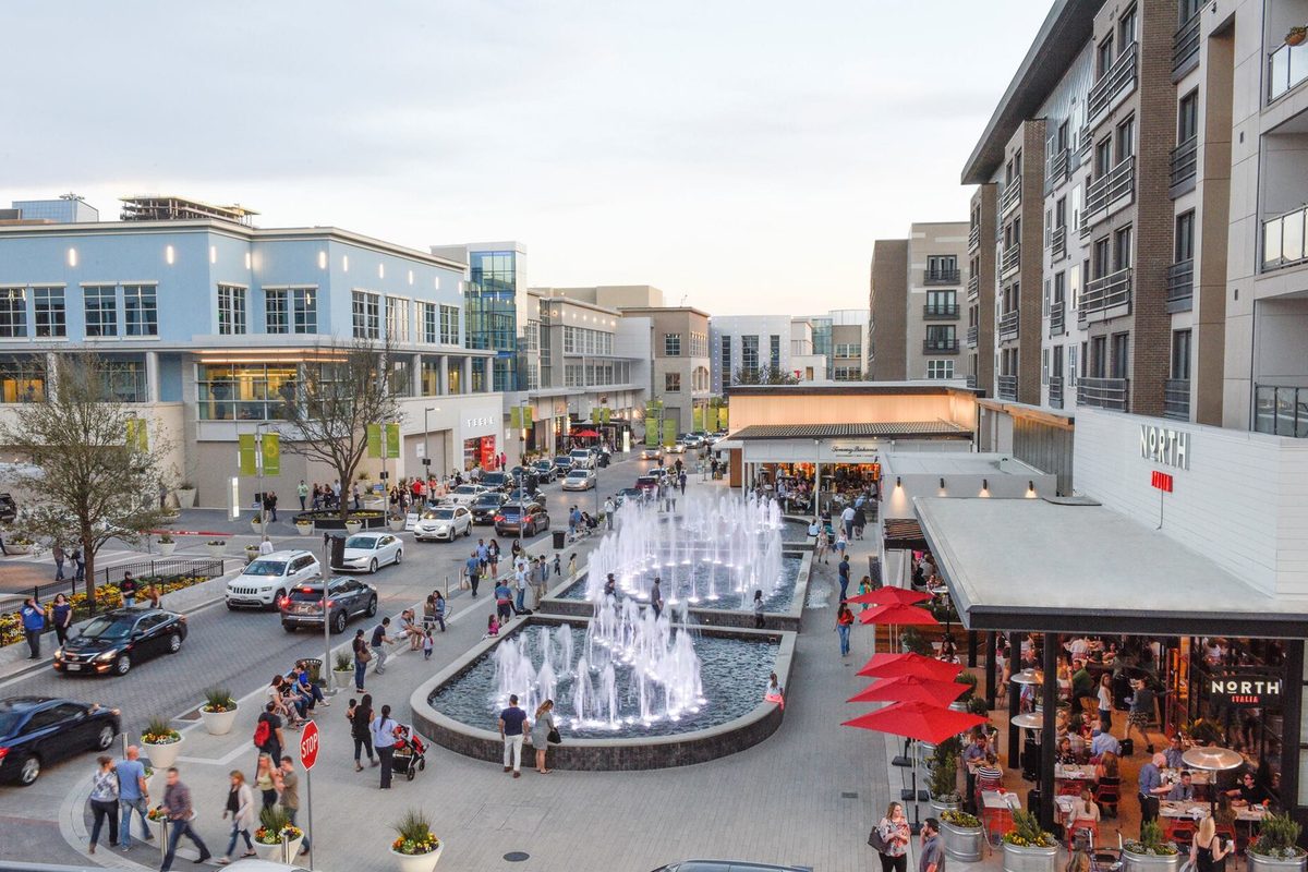 Fountains in the plaza at Legacy West in Plano at dusk, with shoppers, restaurant tables under red umbrellas and cars along the street