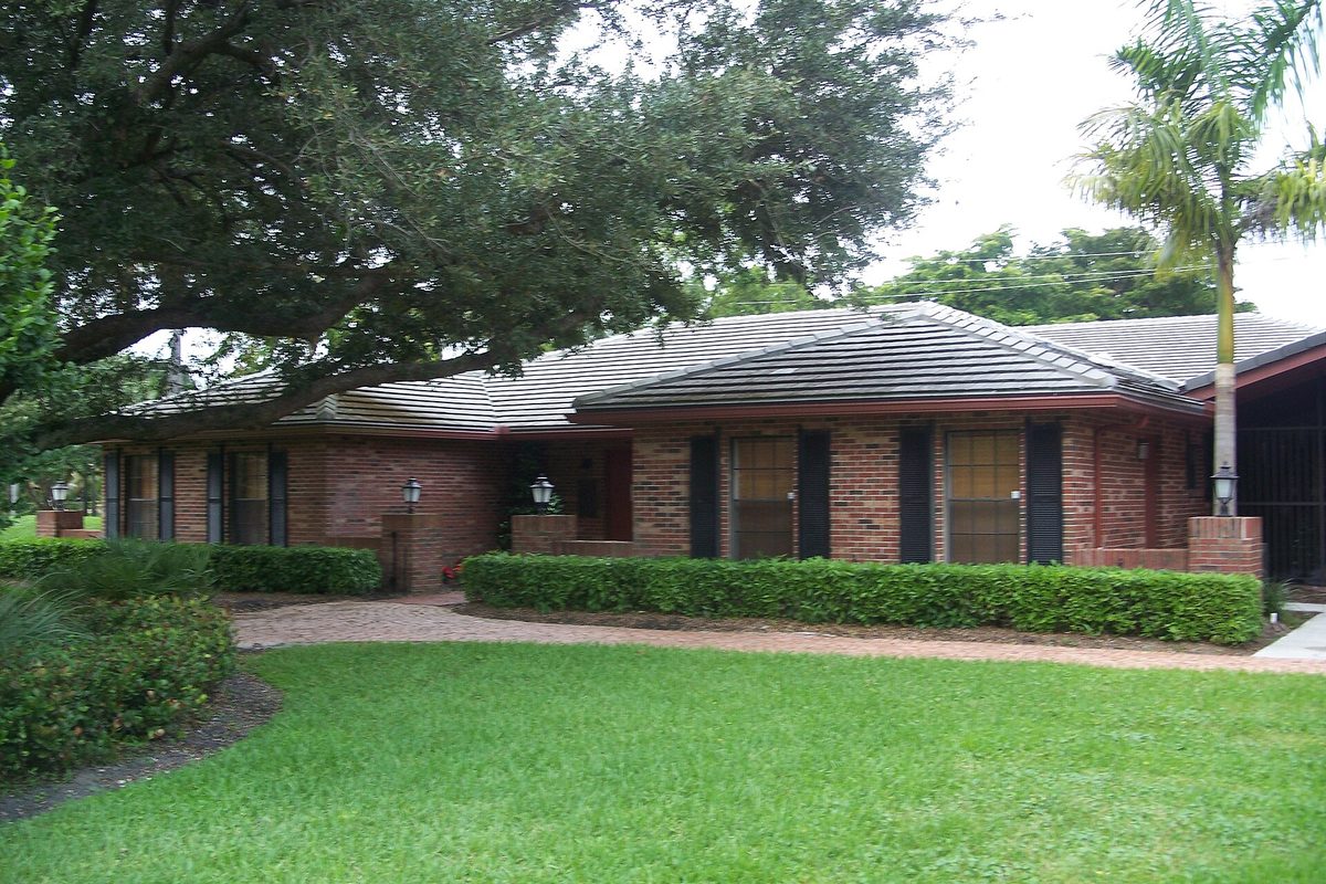 A single-story brick building with a tiled roof, dark shutters, trimmed hedges and a large spreading shade tree over the lawn in Plantation