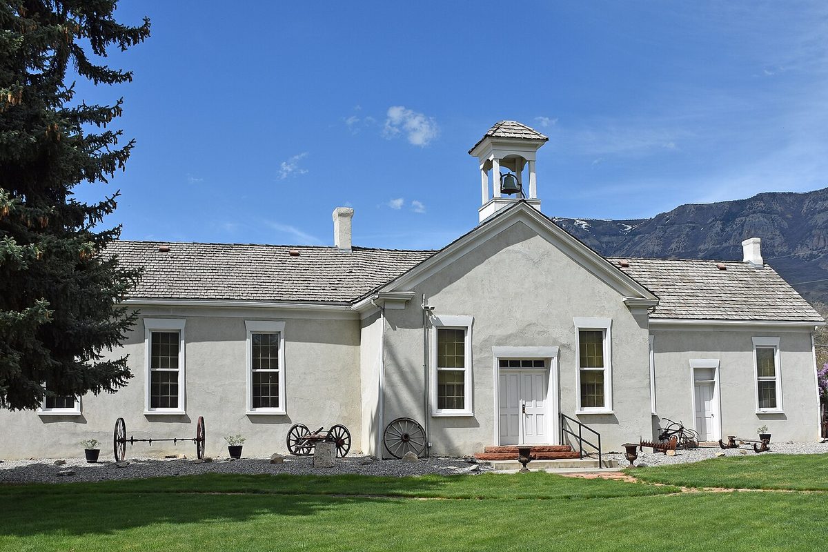 The Old Bell School in Pleasant Grove, a white stucco building with a small bell tower and old wagon wheels along its front, below a rocky mountainside