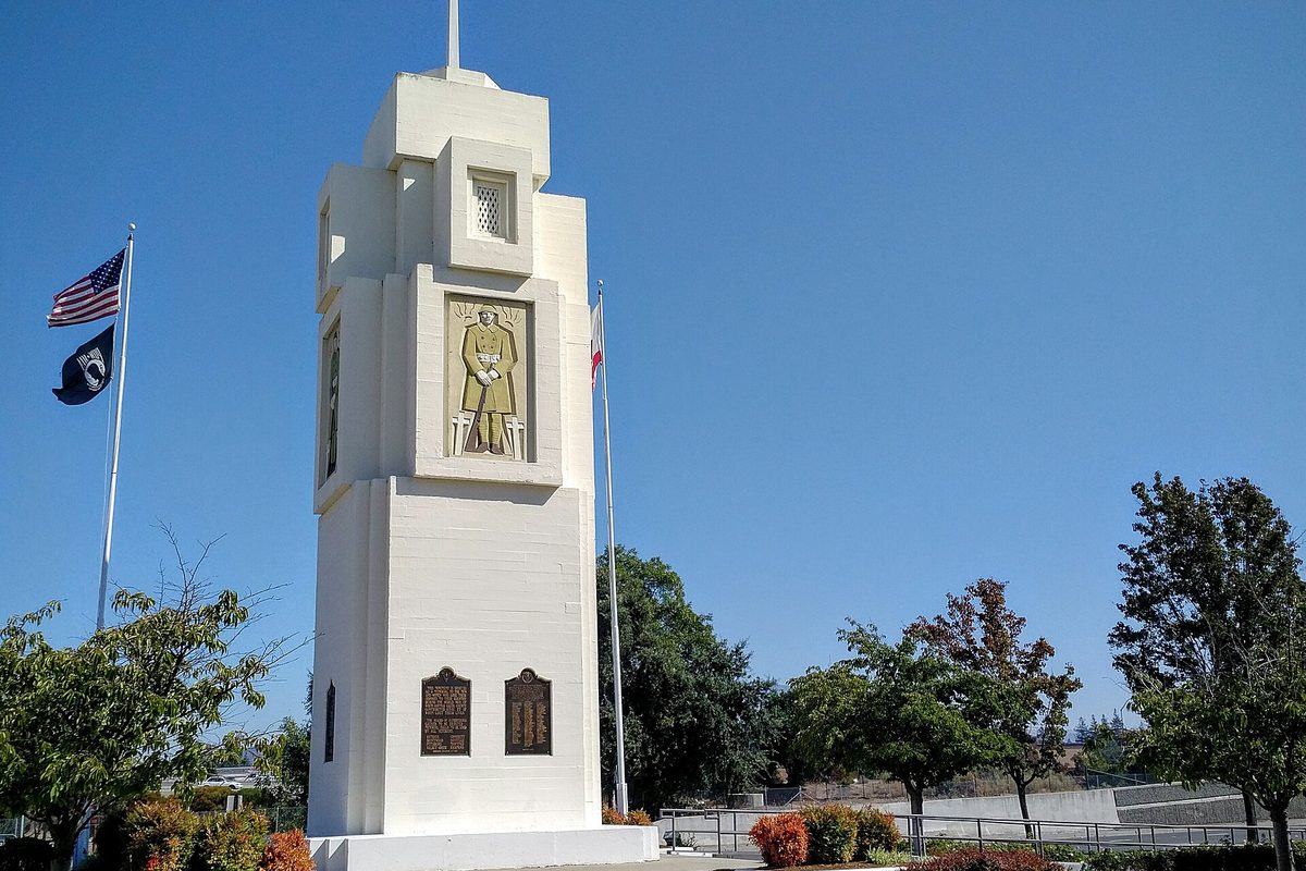 The white Soldiers Memorial tower at Boyd Road in Pleasant Hill, with a carved soldier panel, bronze plaques, and U.S. and POW flags under a blue sky