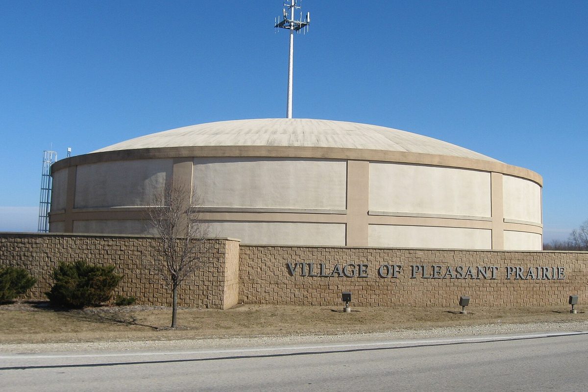 A round, domed village storage building behind a stone wall lettered Village of Pleasant Prairie, under a clear blue sky