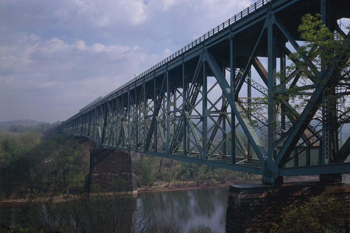 A tall green steel truss railroad bridge on stone piers crossing a wide river, wooded hills on the far bank under a cloudy sky