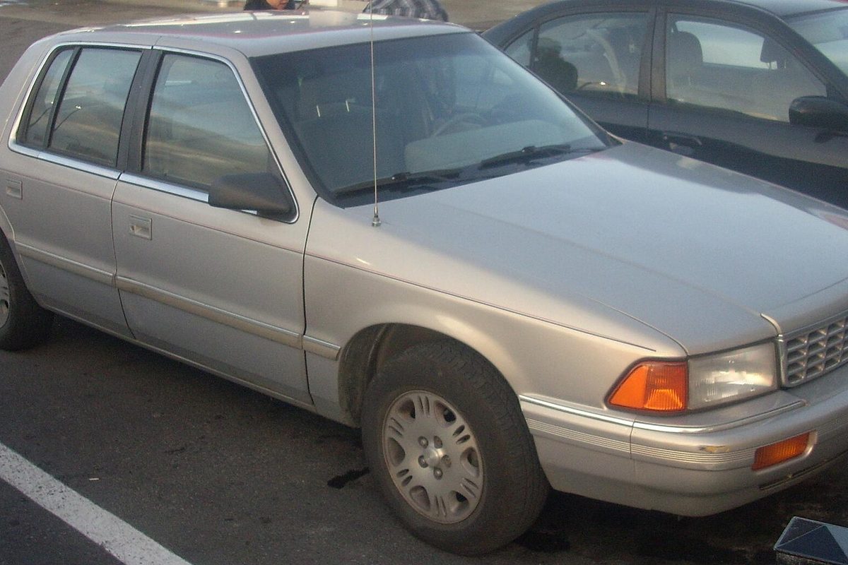 A beige Plymouth Acclaim four-door sedan seen from the front right, parked in a lot next to a dark car