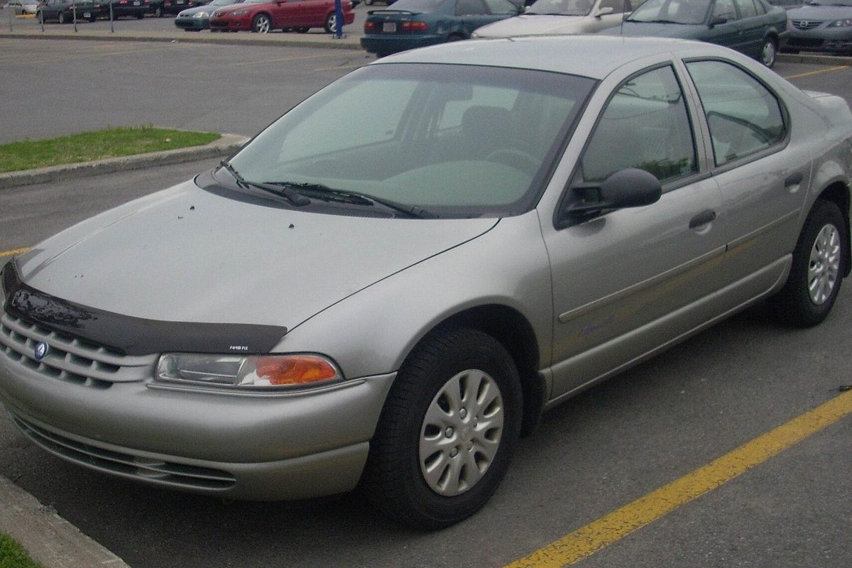 A silver-beige Plymouth Breeze sedan with a black bug deflector on the hood, parked in a lot beside a curb
