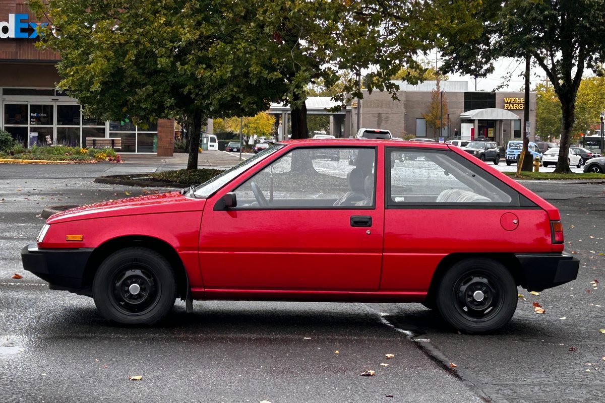A red Plymouth Colt three-door hatchback with black steel wheels, seen from the side in a wet parking lot