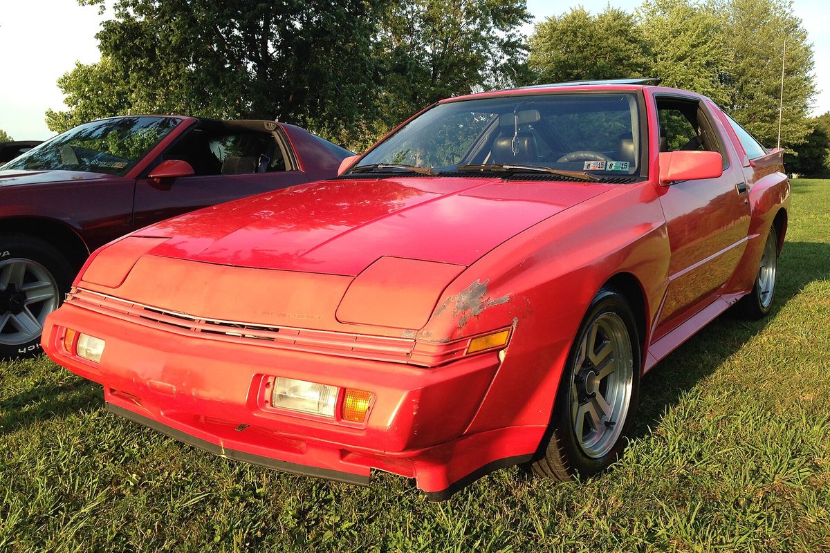 A red Conquest two-door hatchback with flared fenders and peeling paint on its front corner, parked on grass beside other cars