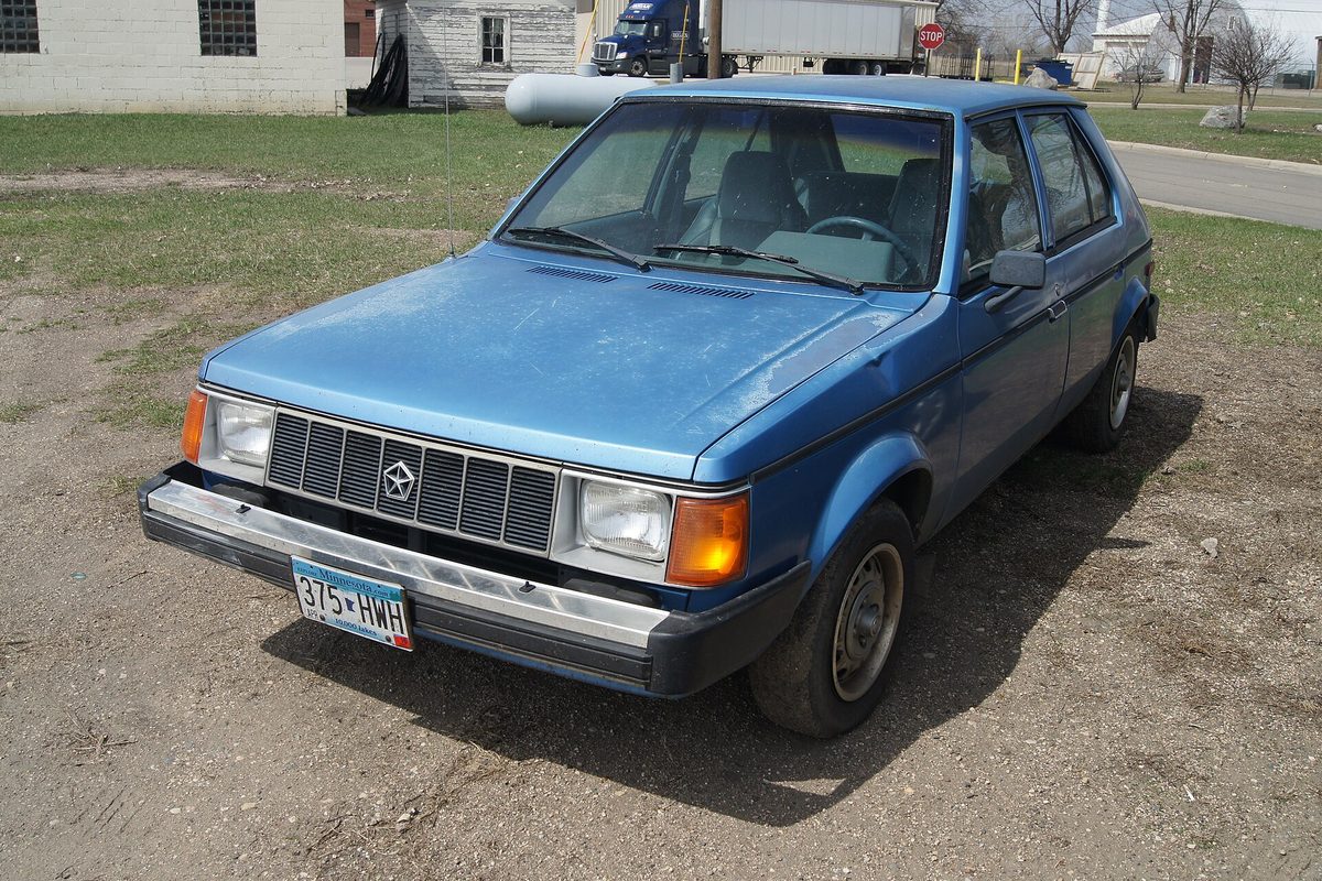 A faded blue Plymouth Horizon hatchback parked on bare dirt next to a patch of grass and an old garage