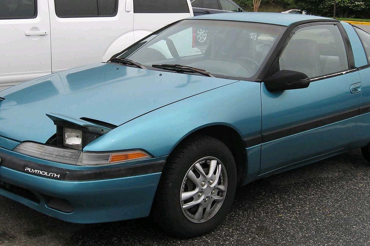 A teal Plymouth Laser hatchback coupe with one pop-up headlamp half raised, parked in a lot beside a white van