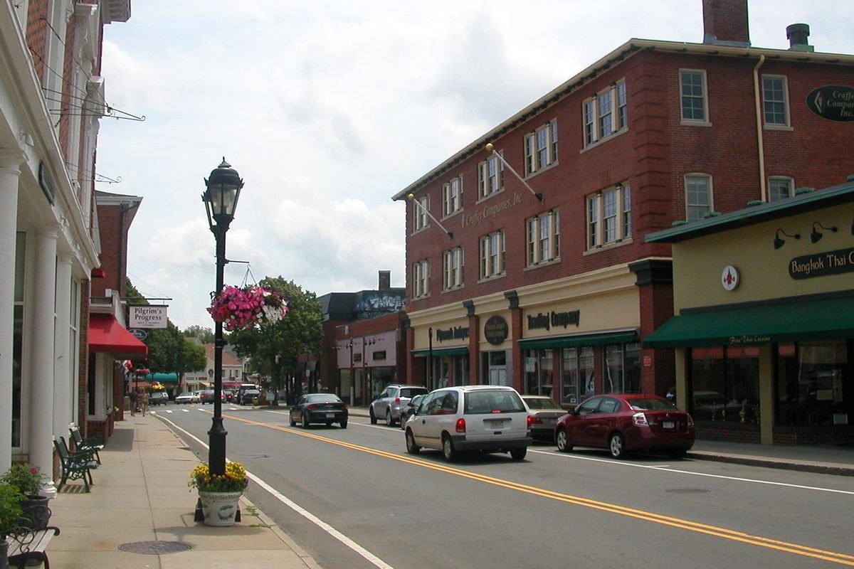 Court Street in downtown Plymouth, with a black lamppost holding a pink flower basket, brick storefronts and cars driving toward Town Brook