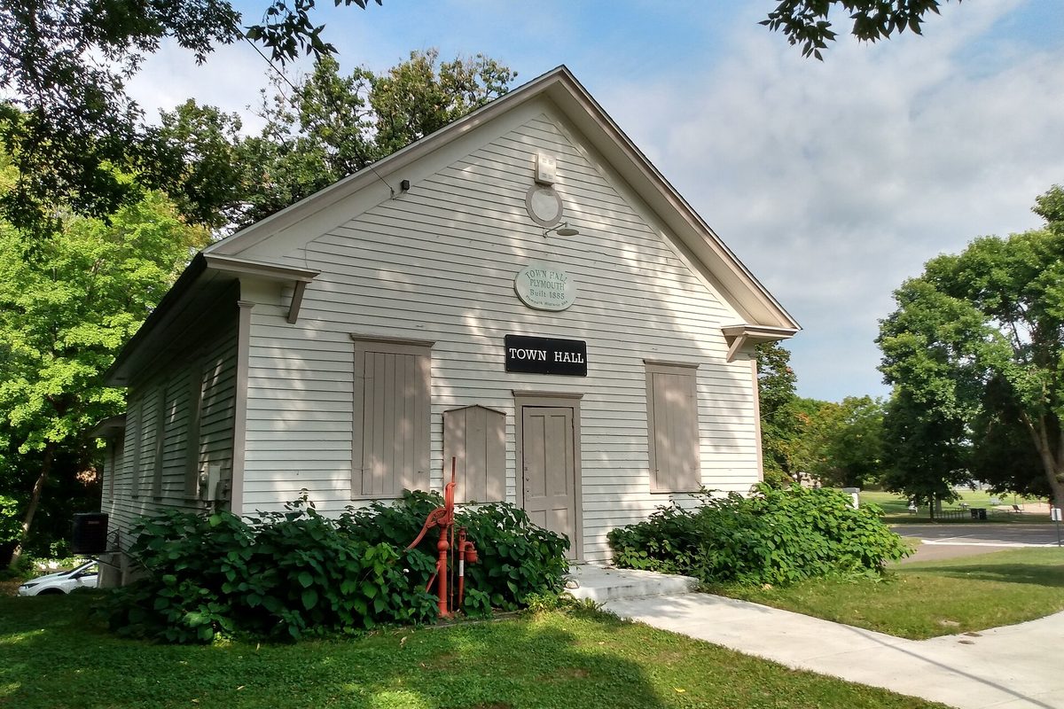 A small white clapboard building with a Town Hall sign over its door, a red hand pump in front and green shrubs around it