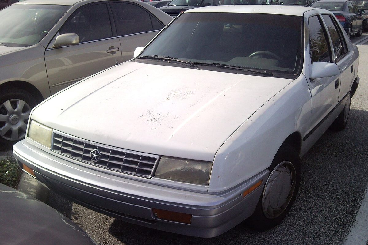 A white Plymouth Sundance hatchback with sun-damaged paint on the hood, parked between two cars in a lot