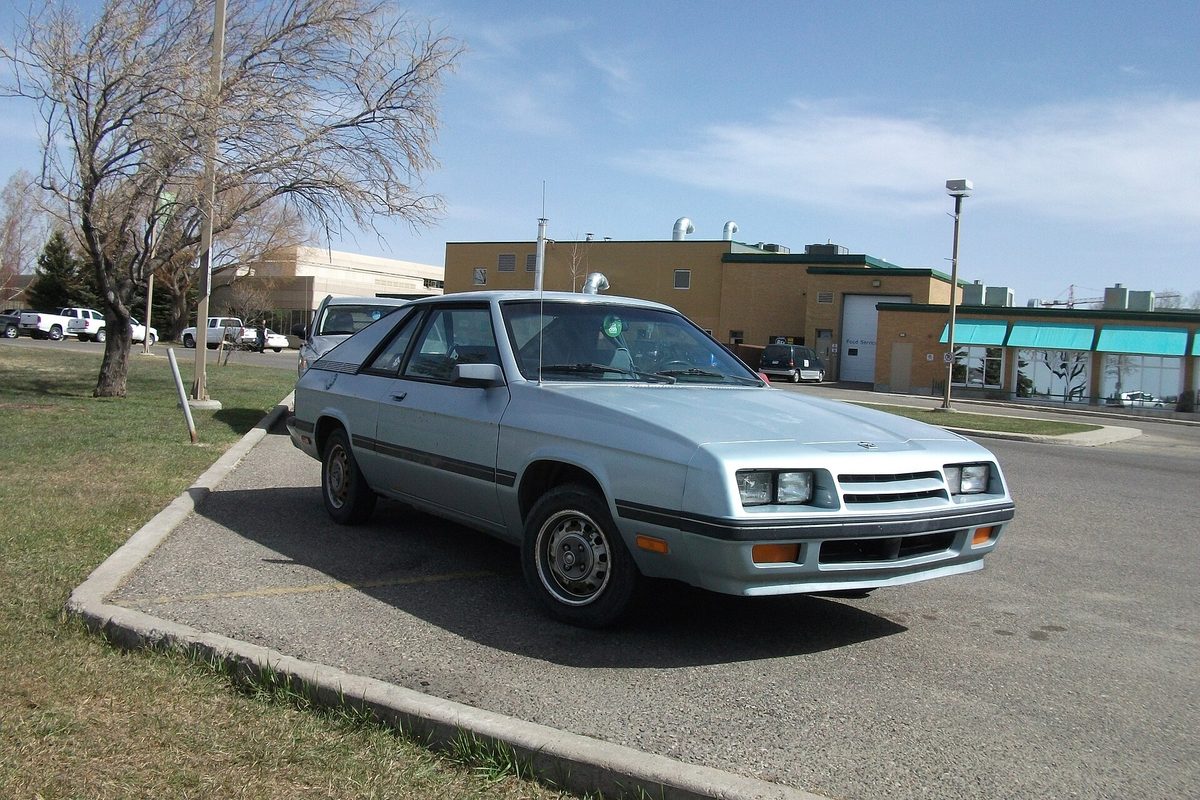 A pale silver-blue Plymouth Turismo hatchback coupe parked in a lot beside a bare tree on a clear day