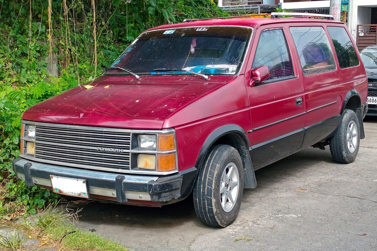 A faded red first-generation Plymouth Voyager minivan with a scuffed grille and bumper, parked on a cracked street beside overgrown plants
