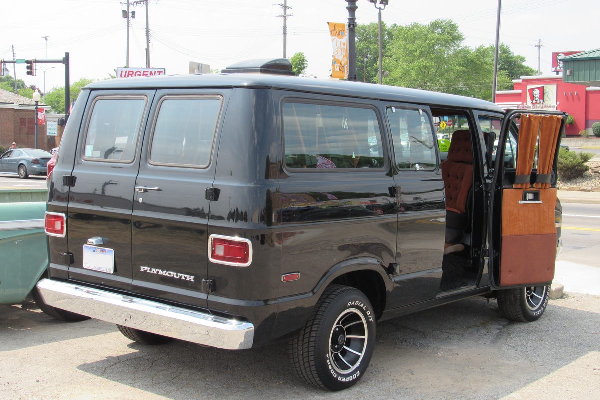 A black 1974-1977 Plymouth Voyager van seen from the rear corner, its side doors open to a brown interior, parked on a lot in Ohio