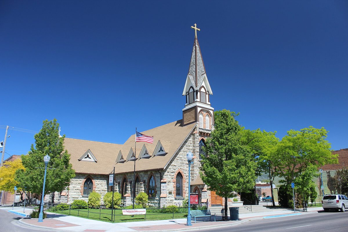 Trinity Episcopal Church in Pocatello, a stone church with a tall steeple and gold cross, an American flag and green trees under a deep blue sky