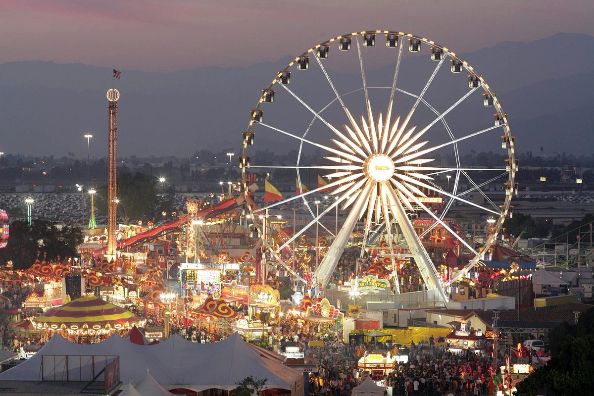 The Los Angeles County Fairgrounds in Pomona at dusk, with a lit Ferris wheel, a tall ride tower, striped tents and crowds below the hills