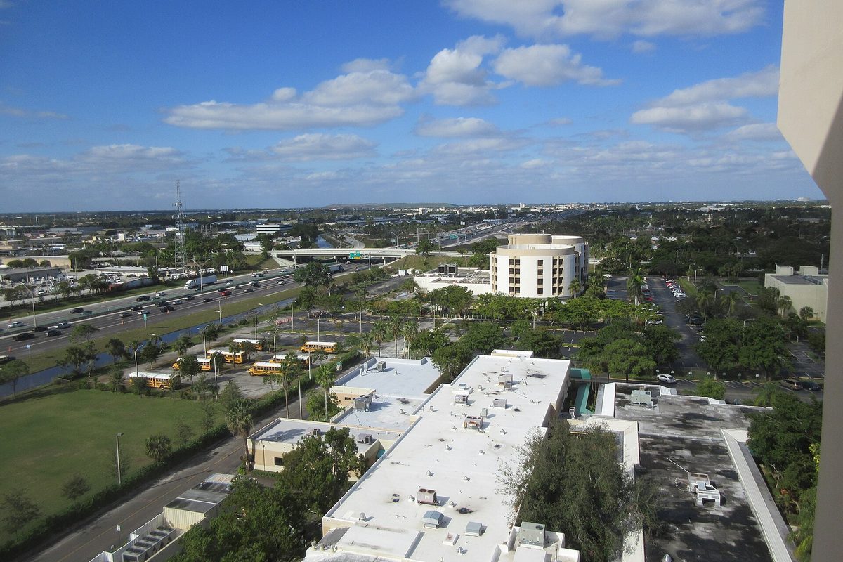 View from a high-rise over Pompano Beach: flat white rooftops, yellow school buses in a lot, a busy highway with an overpass and a round tower among trees