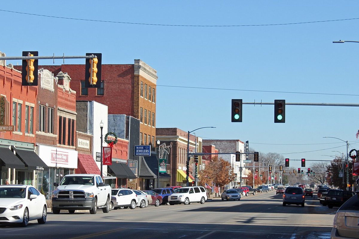 East Grand Avenue in downtown Ponca City, with two- and three-story brick storefronts, cars parked along the curb and traffic lights over the street under a clear blue sky