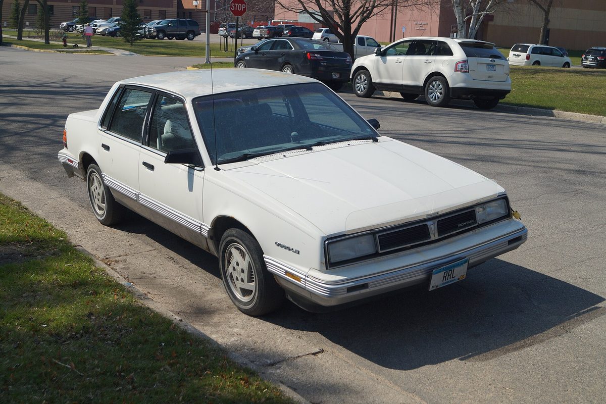 A white 1988 Pontiac 6000 LE four-door sedan with silver lower body trim, parked at a curb with a lawn in front