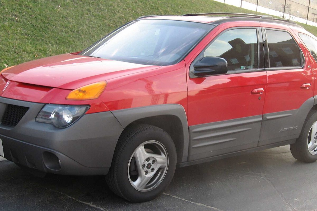 A red Pontiac Aztek with gray lower body cladding, parked at the foot of a grassy slope