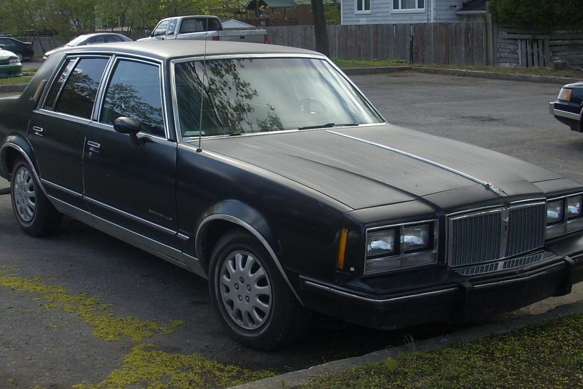 A black 1984-1986 Pontiac Bonneville four-door sedan with faded paint and plain hubcaps, parked on a residential street