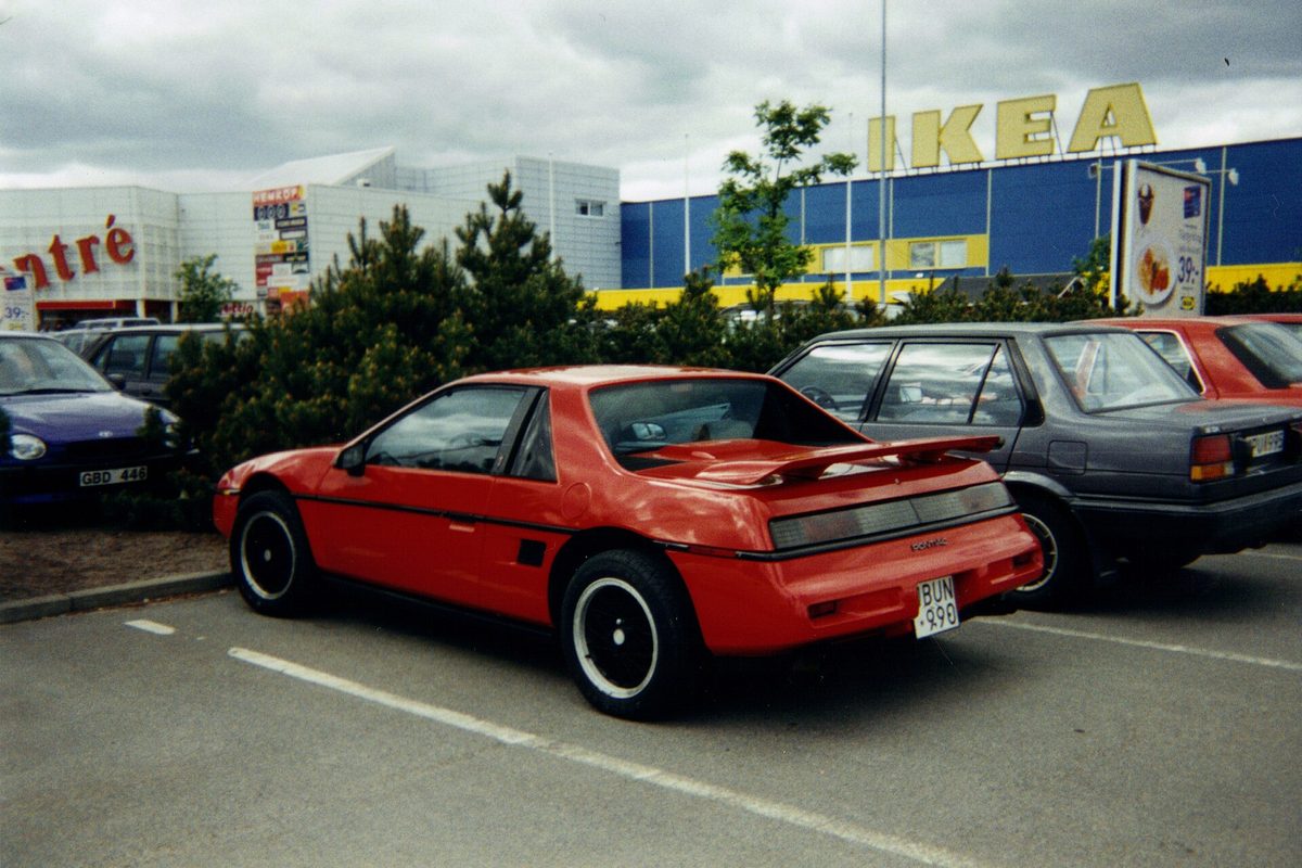 A red Pontiac Fiero coupe with a rear spoiler and black wheels, parked in a store lot beside other cars