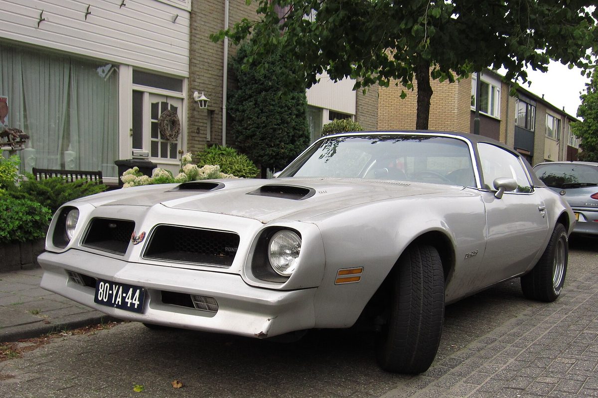 A silver Pontiac Firebird Formula with twin hood scoops, parked on a brick street in front of row houses