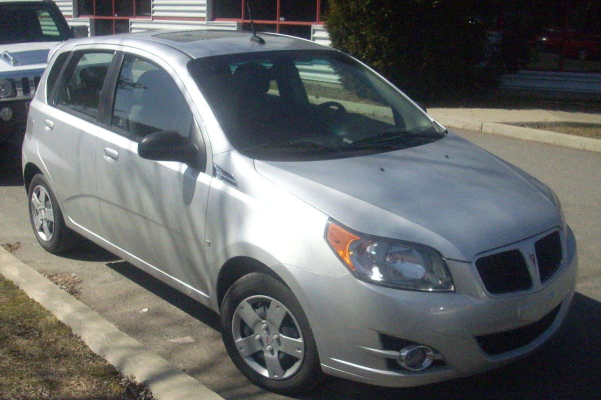 A silver 2010 Pontiac G3 five-door hatchback parked at a curb beside a building, with a truck behind it