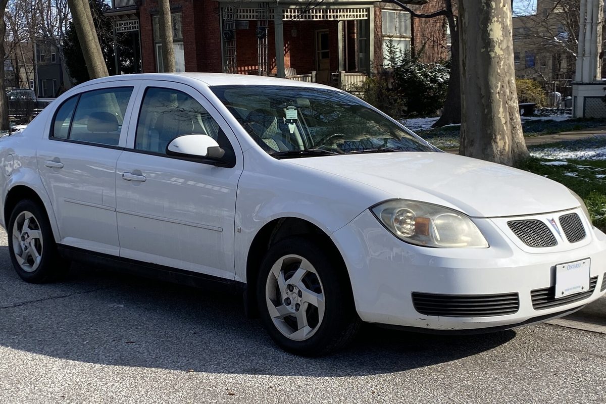 A white Pontiac G5 sedan parked at a curb on a residential street, with patches of snow on the lawn behind it